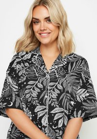 Svart kortärmad pyjamaströja med vita bladmönster, knappar och krage, tillverkad av lätt material för ökad komfort.