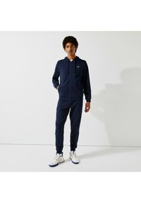 Haut de survêtement zippé bleu marine avec poches kangourou et joggers assortis ; baskets blanches avec des accents rouges et bleus ; texture douce, coupe athlétique.