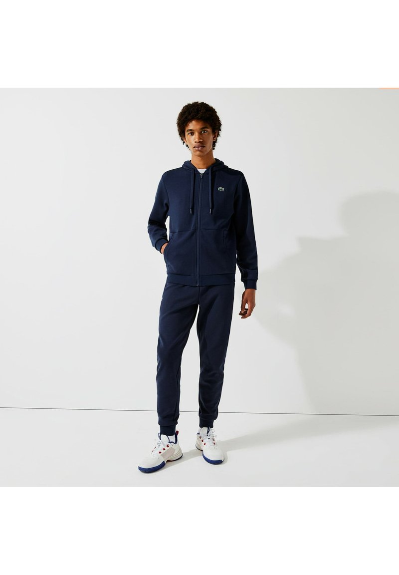 Haut de survêtement zippé bleu marine avec poches kangourou et joggers assortis ; baskets blanches avec des accents rouges et bleus ; texture douce, coupe athlétique.