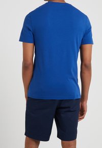 Muž oblečený v jednoduchém modrém tričku s krátkým rukávem a tmavě modrých šortkách, stojící zády k jednoduchému bílému pozadí.