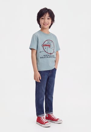 Garçon souriant debout les mains dans les poches, portant un t-shirt bleu Stella McCartney, un jean foncé et des baskets montantes rouges sur fond blanc.