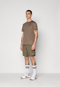 T-shirt marron à manches courtes, shorts vert olive, baskets blanches avec semelles en caoutchouc, et chaussettes blanches montantes avec rayures noires.