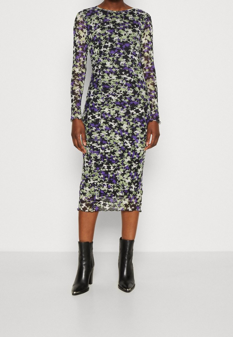 Femme portant une robe midi ajustée à manches longues avec un motif floral noir, violet et vert, associée à des bottines noires à talons.