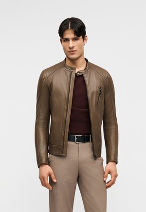 Mees, kes kannab pruuni nahast lukuga jope tumepunase kampsuni, beežide pükste ja musta vööga, seisab ühevärvilise tausta ees.