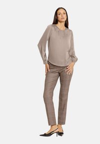 Beige Bluse mit langen Ärmeln und Raffung am Ausschnitt, kombiniert mit braunen Karohosen und schwarzen spitz zulaufenden Absatzschuhen.