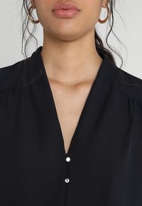 Blusa nera con scollatura a V profonda, pieghe sottili sulle spalle e tre bottoni metallici visibili sul colletto. Tessuto con texture liscia.