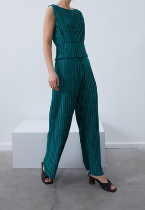 Vrouw draagt mouwloze teal geplooide top en bijpassende brede pantalons met zwarte sandalen met hak, staand op een grijze vloer bij een wit blok.