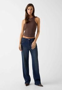 Bruine geribbelde mouwloze top gecombineerd met donkerblauwe jeans met wijde pijpen. De jeans hebben vijf zakken en een knoopsluiting. Puntige schoenen maken de look af.