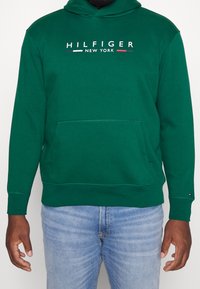Grøn hættetrøje lavet af blødt stof, med en frontlomme og logo-tryk "HILFIGER NEW YORK" i hvidt med farvede accenter.