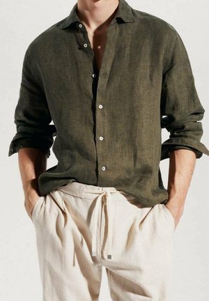 Mand iført en olivengrøn skjorte med knapper, ærmerne rullet op, og lysebeige bukser med snøre, hænderne i lommerne.