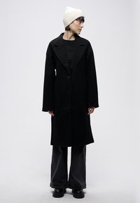 Melns garš mētelis ar atvērtu apkakli, pogu aizdares un platiem piedurknēm. Valkāts pār melnu topu, kombinēts ar brīvā stila tumšām biksēm un melniem apaviem.