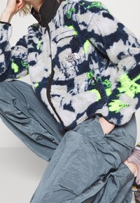 Kurtka polarowa w kolorach niebieskim, szarym i neonowo zielonym, z zamkiem z przodu, wysokim kołnierzem i naszywką z logo. Do kompletu srebrne, luźne spodnie.