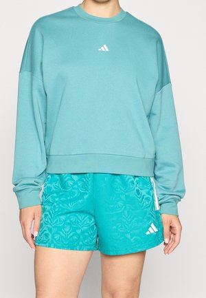 Person iført turkis langærmet sweatshirt og turkis mønstrede atletikshorts med hvidt logo på shorts og sweatshirt.