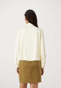 Blusa color crema con maniche lunghe e leggermente gonfiate e colletto; texture liscia, senza fantasie, abbinata a una gonna verde oliva scura.