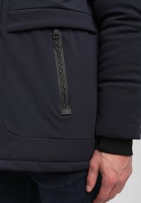 Tumši zils piepūsts mētelis ar sānu rāvējslēdzēja kabatu, teksturētu audumu un melniem ribu aprocēm. Gluda, eleganta dizaina forma ar tīrām līnijām.