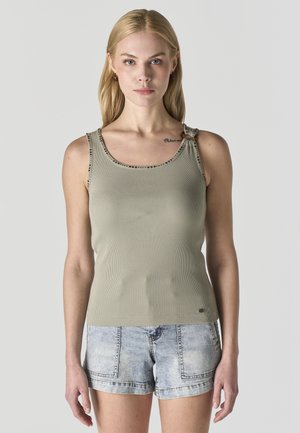 Mujer rubia con una blusa sin mangas de color verde oliva con tirante anudado y shorts de mezclilla claros, de pie frente a un fondo liso.