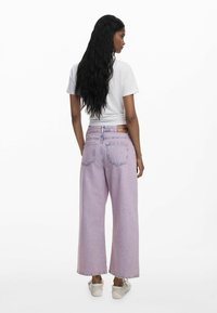 Vaqueros de mezclilla morados de pernera ancha con cintura alta, bolsillos traseros y etiqueta de cuero en la cintura. Combinados con una camiseta blanca y zapatillas.