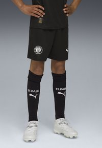 Svarta fotbollsshorts med en cirkulär FC St. Pauli-logga, kombinerat med knähöga svarta strumpor som visar "ST. PAULI" i vitt. Silverfärgade fotbollsskor med texturerad design.
