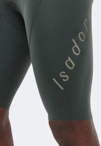 Κοντινή λήψη σκούρων πράσινων συμπιεστικών σορτς με κάθετη ανακλαστική επιγραφή στο μηρό που γράφει "Isadore."