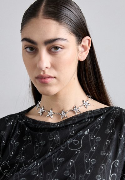 Silver star-shaped necklace made of metal, featuring a linked design. Worn over a black top with a subtle pattern of safety pins.