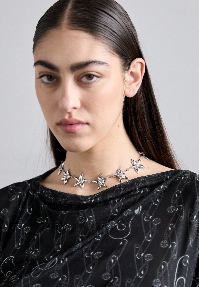 Sternförmige Halskette aus Metall in Silber, mit einem verlinkten Design. Getragen über einem schwarzen Oberteil mit dezentem Muster von Sicherheitsnadeln.