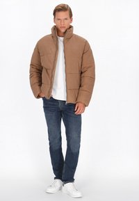 Bruine gewatteerde jas met hoge kraag, geborduurd patroon en ritssluiting, gecombineerd met een wit T-shirt, blauwe jeans en witte sneakers.