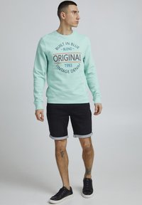Mentazöld pulóver "Eredeti Vintage Denim" grafikával, fekete farmer sorttal és fekete sportcipővel párosítva. Egyszerű design, mandzsettákkal.