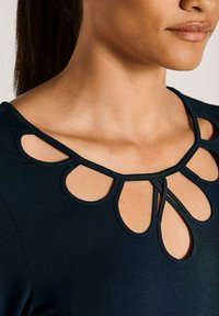 Frau, die ein schwarzes Oberteil mit tränenförmigen Ausschnitten am Ausschnitt trägt und ein natürliches Make-up auf Lippen und Haut hat.