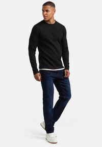 Camiseta negra de manga larga con textura acanalada, superpuesta sobre una camiseta blanca de cuello redondo, combinada con jeans azul oscuro y zapatillas blancas. Diseño simple y limpio.