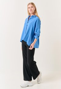 Chemise oversize bleu clair à boutons et col, associée à un pantalon large noir et des baskets blanches. Texture lisse et coupe décontractée.