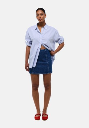 Chemise oversized rayée bleu et blanche, minijupe en denim, ballerines rouges avec des nœuds, offrant une coupe décontractée et un style casual.