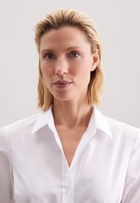 Biała koszula zapinana na guziki z klasycznym kołnierzykiem, o gładkiej fakturze i lekko dopasowanym kroju. Jasne, przejrzyste tło podkreśla detale.