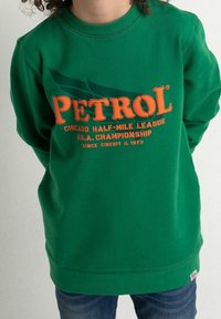 Barn som bär en grön sweatshirt med orange "PETROL" text och mindre texten "Chicago Half-Mile League U.S.A. Championship Since Circuit IL 1973".