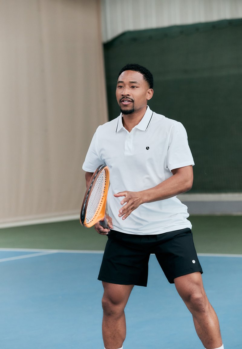 Weißes Polohemd mit schwarzem Besatz, kombiniert mit schwarzen Shorts. Hält ein oranges Tennisschläger auf einem blauen Tennisplatz.