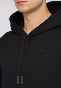 Czarna bluza z kapturem wykonana z teksturowanej tkaniny. Posiada przednią kieszeń kangurką, sznurki z metalowymi końcówkami oraz małe logo na klatce piersiowej.