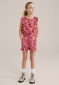 Kratka roza kombinezon z nabrano bluzo in abstraktnim vzorcem. Ima žepne stranske zadrge, prilagojeno pas s trakom in naborke na kratkih hlačah. Nosite ga z bež škornji.