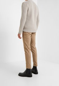 Mężczyzna ubrany w beżowy sweter dzianinowy, khaki spodnie i czarne skórzane botki Chelsea stoi plecami do białego tła.