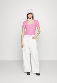 Top rosa de mangas curtas com decote quadrado, combinado com calças brancas de perna larga e um boné lilás claro. Acessorizado com um lenço.