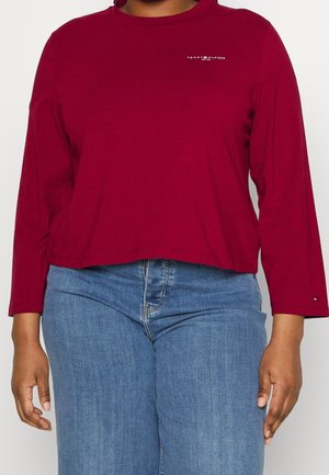 Person trägt ein burgunderrotes Langarmshirt von Tommy Hilfiger und blaue Jeans, steht vor einem schlichten hellen Hintergrund.