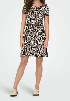 Femme portant une robe courte à manches courtes, longueur genou, imprimé animal noir et beige, avec des mocassins beiges, se tenant devant un fond uni.