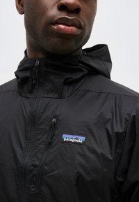 Κοντινό πλάνο ενός ατόμου που φοράει μαύρο μπουφάν Patagonia με φερμουάρ στο στήθος και κουκούλα, εμφανίζοντας το λογότυπο της μάρκας.
