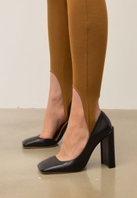 Črne usnjene pete z kvadratnimi nosovi in čvrstimi petami, v kombinaciji z oprijetimi bež legicami, ki imajo izreze nad gležnji.