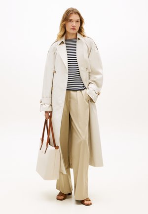 Woman wearing beige trench coat, striped black and white shirt, wide beige pants, brown sandals, holding large beige tote bag with brown handles.
