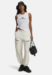 Žena v bílém bezrukávovém tričku Under Armour, světle šedých cargo kalhotách, černých teniskách a klobouku typu bucket, držící černý stahovací batoh.