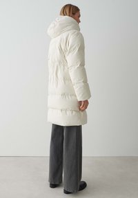 Parka blanche longue et bouffante avec capuche, dotée d'une taille froncée et de larges manches. Associée à un pantalon noir à jambes larges et des bottes noires.