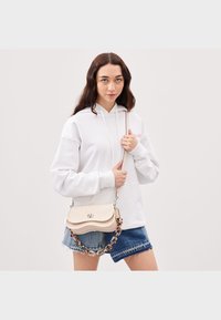 Biała bluza z kapturem ze sznurkami, zestawiona z niebieskimi szortami dżinsowymi. Modelka trzyma kremową torebkę na łańcuszku, ozdobioną falistym wzorem.