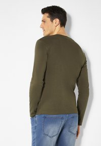 Langärmliges Shirt aus olivgrünem Stoff, runder Ausschnitt, schlanker Schnitt, glatte Textur, kombiniert mit hellblauen Jeans mit Gesäßtaschen.