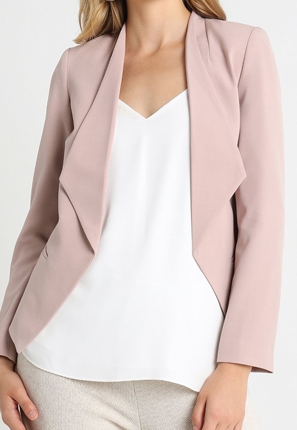 Femme portant un blazer rose clair sur un haut blanc à col en V et un pantalon texturé de couleur claire, vue des épaules aux hanches.