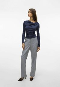 Maglietta navy a maniche lunghe con collo drappeggiato, abbinata a pantaloni a quadri grigio chiaro e scarpe con tacco marroni con dettagli in metallo decorativo.
