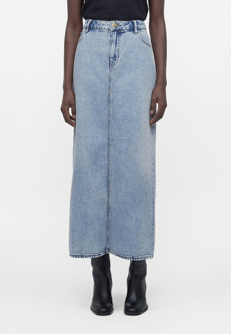 Γυναίκα που φοράει μια φούστα maxi από ανοιχτό μπλε τζιν με ψηλή μέση, ραφή μπροστά και μαύρα μποτάκια αστραγάλου σε λευκό φόντο.